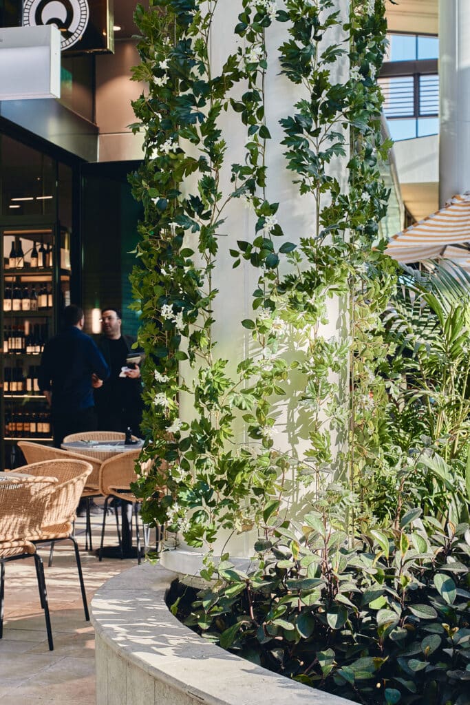Leafy indoor café area with vines climbing a column, wicker chairs, and two people talking near a wine display.