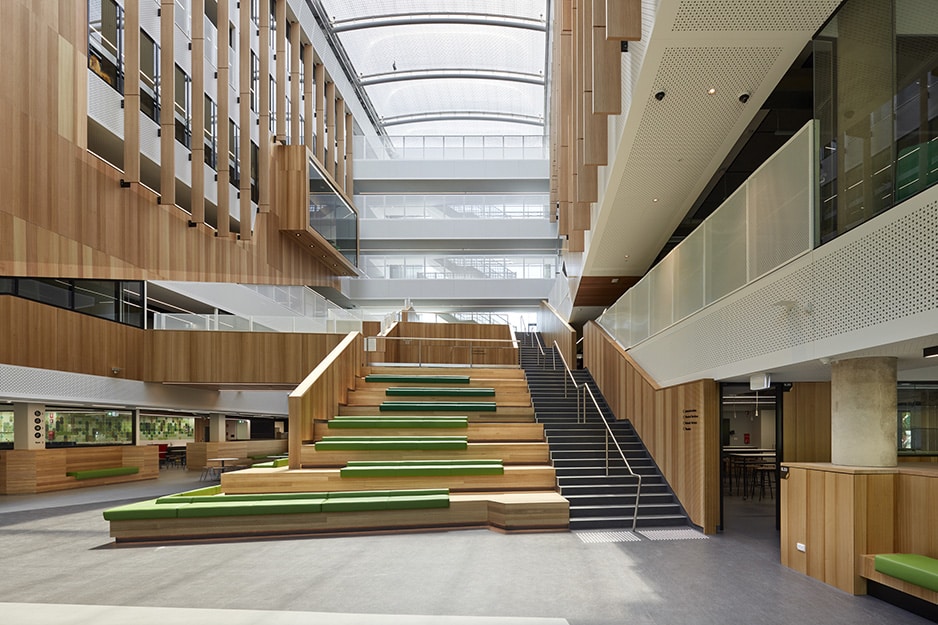 Modern multi-level atrium with timber finishes, tiered seating, open staircases, and a large skylight ceiling.