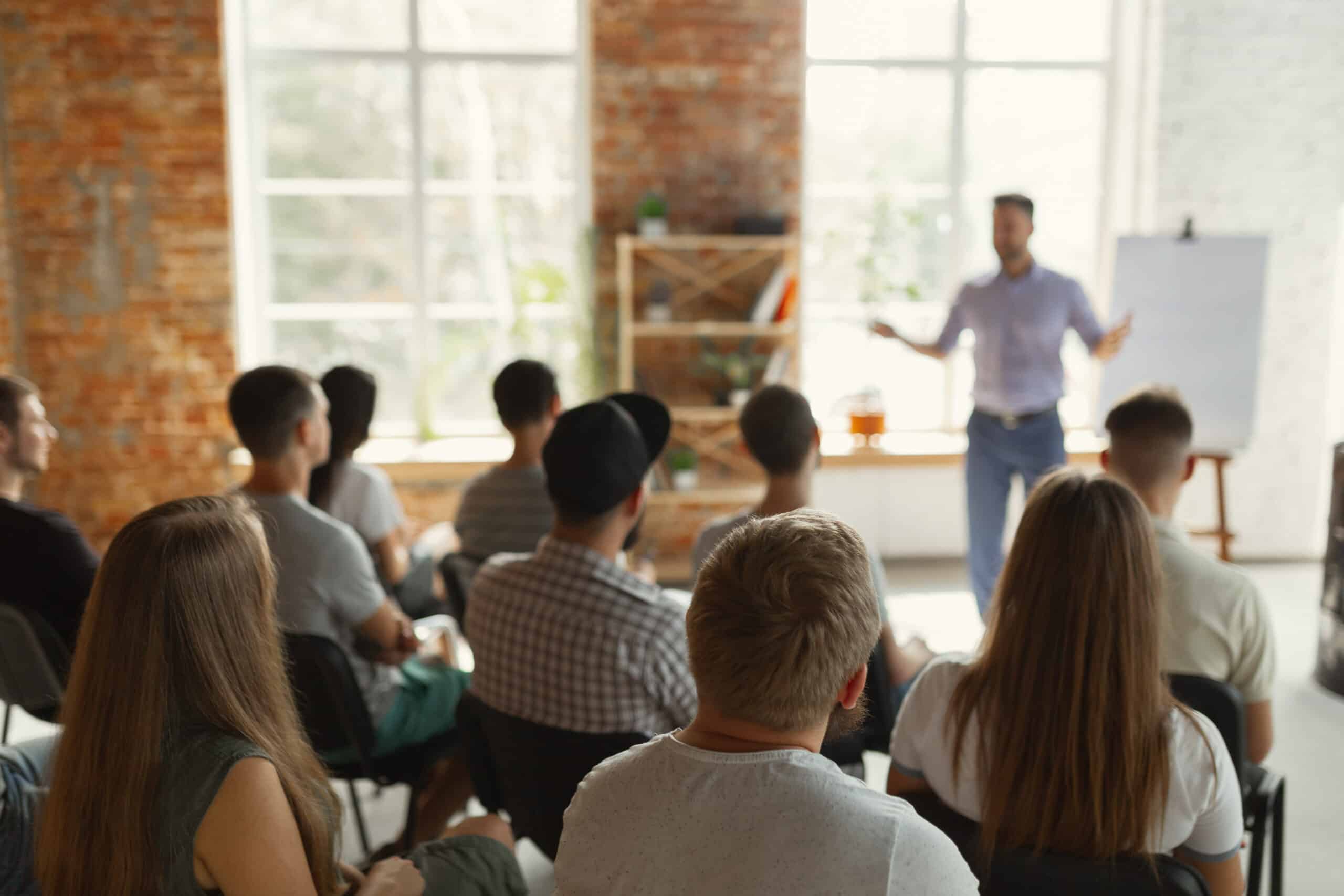 Male speaker giving presentation in hall at workshop. Audience or conference hall. Rear view of unrecognized participants in audience. Education concept.