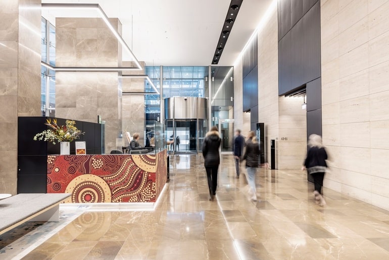 Contemporary office lobby with polished stone finishes, reception desk artwork, glass entryways, and people walking through the space.