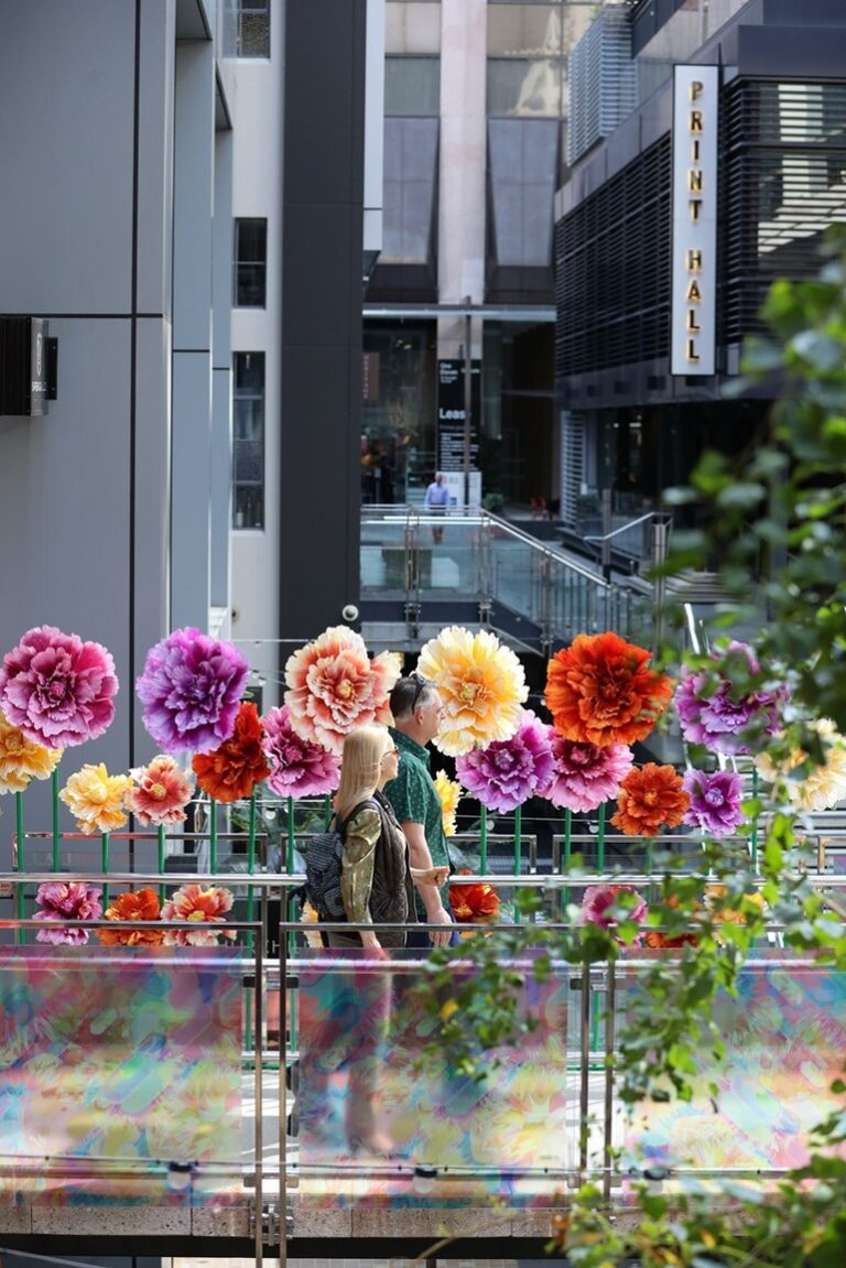 People walking through a modern urban walkway decorated with colourful oversized floral installations between contemporary office buildings.
