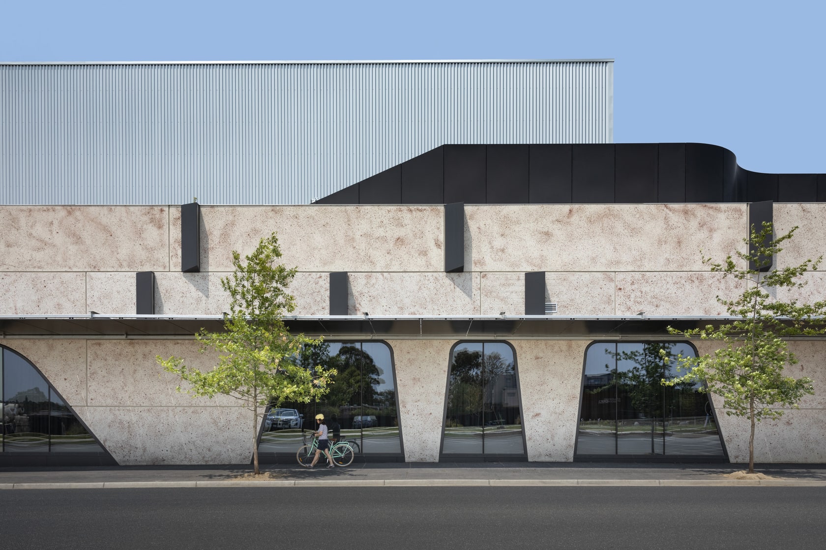 Contemporary commercial building with textured stone cladding, curved black architectural elements, corrugated metal upper façade, and large irregularly shaped windows facing a tree-lined street.