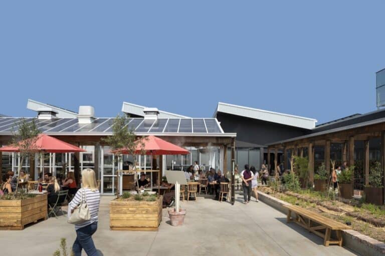 Outdoor dining courtyard with timber planters, red umbrellas, landscaped garden beds, and people gathering between contemporary pavilion-style buildings under a clear blue sky.