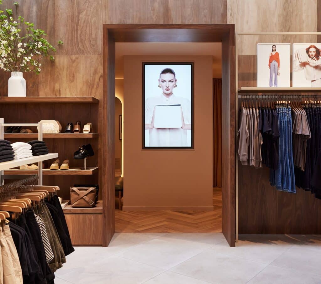 Modern retail interior with warm timber finishes, neatly arranged clothing racks and shelves, and a central doorway featuring a digital display of a model holding a tablet.