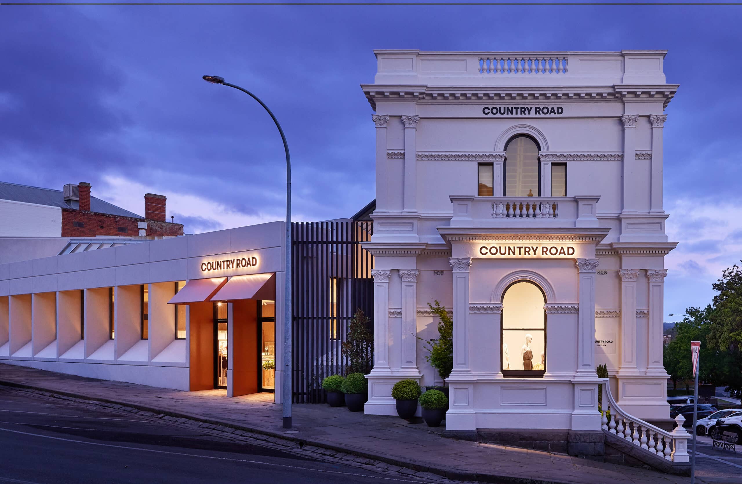 Country Road store in a restored heritage building at dusk, featuring a white classical façade with arched windows, modern extension to the left, and warm interior lighting on a quiet street corner.