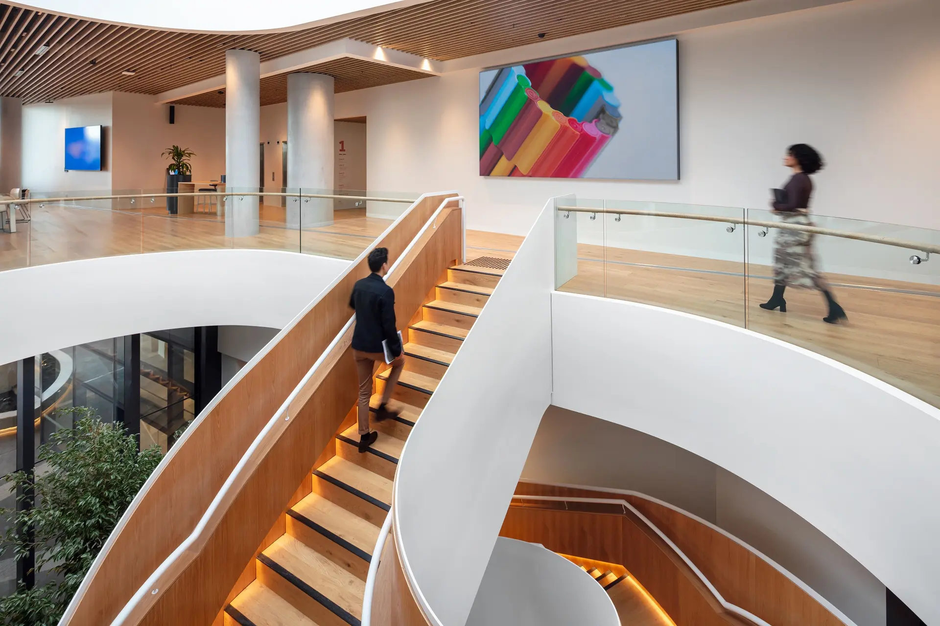Modern interior with a curved staircase, wood finishes, glass railings, and people walking past colorful wall art.