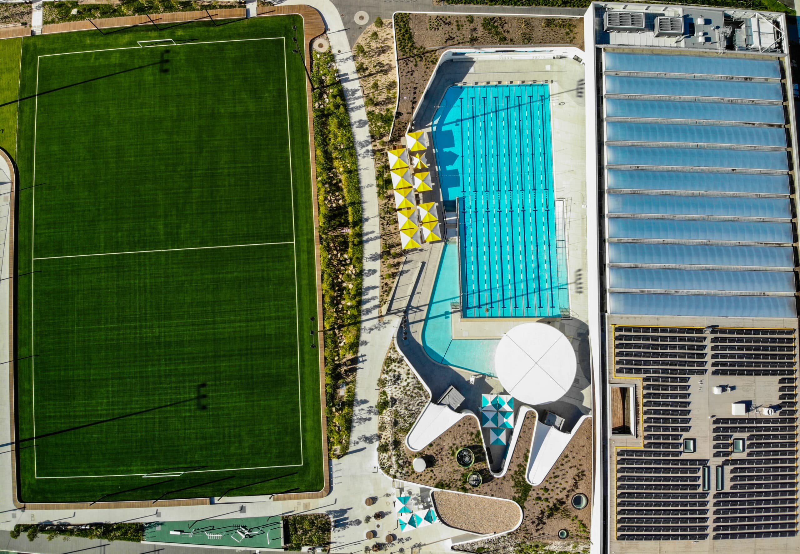 Aerial view of sports field, outdoor swimming pool, and aquatic centre complex with solar panels and landscaped walkways.