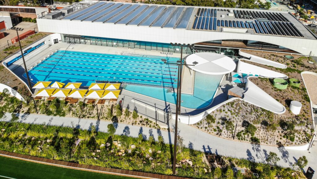 Aerial view of sports field, outdoor swimming pool, and aquatic centre complex with solar panels and landscaped walkways.