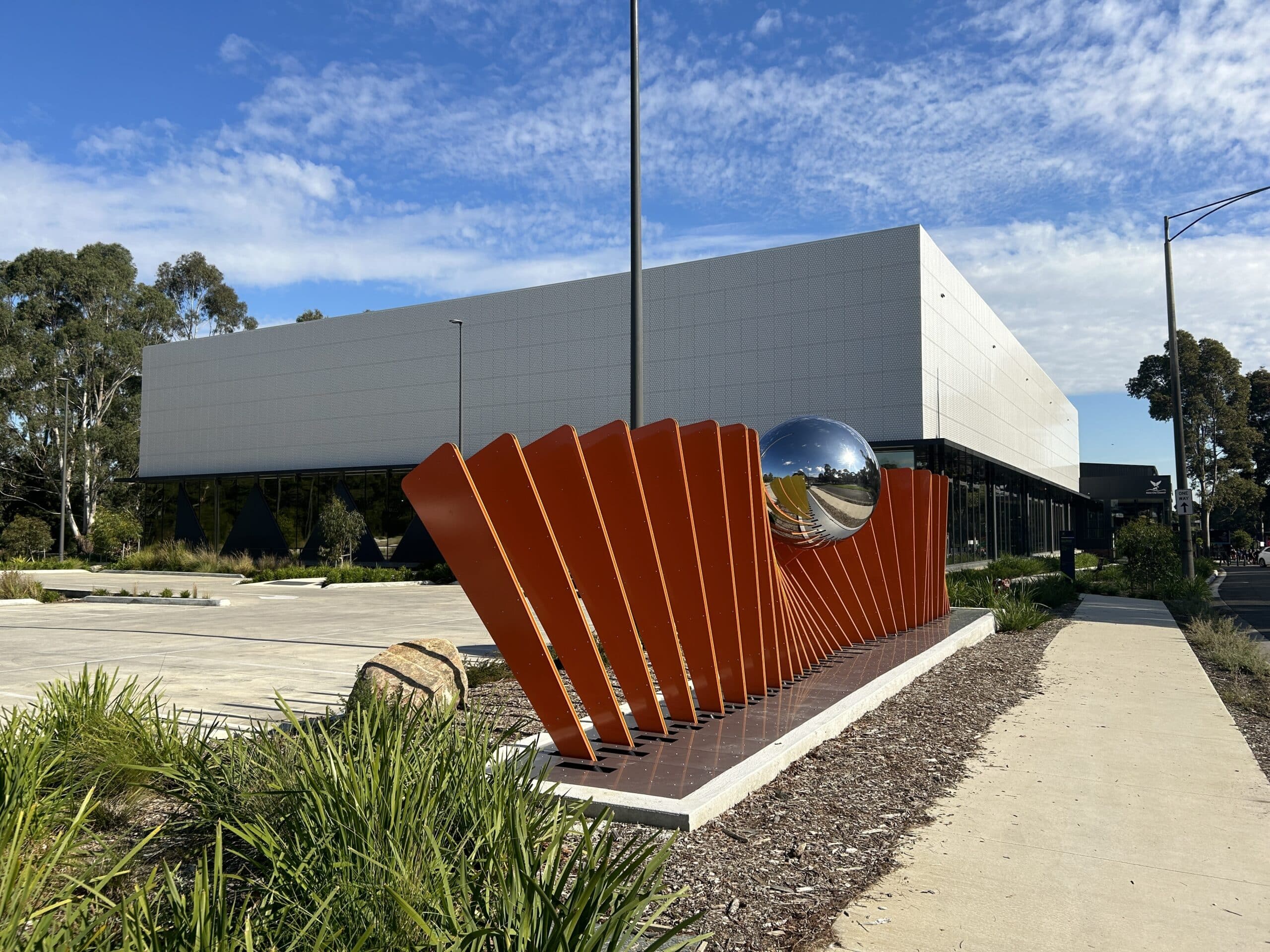 Modern sports facility exterior with a white geometric façade, featuring a red sculptural installation with angled panels and a reflective sphere, set beside a landscaped walkway under a blue sky.