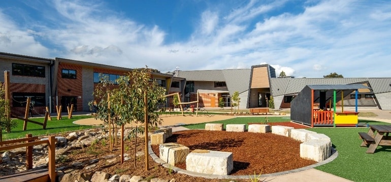 Contemporary school campus with landscaped playground, outdoor seating, and modern classroom buildings under a blue sky.