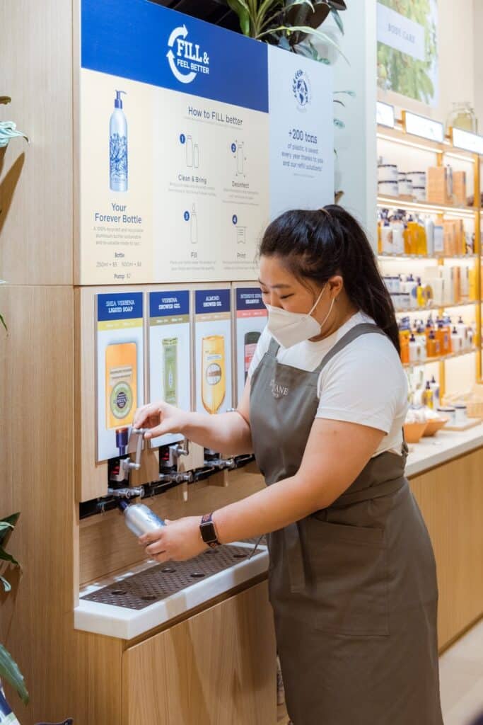 Retail staff member refilling a reusable bottle at L’Occitane in-store refill station.