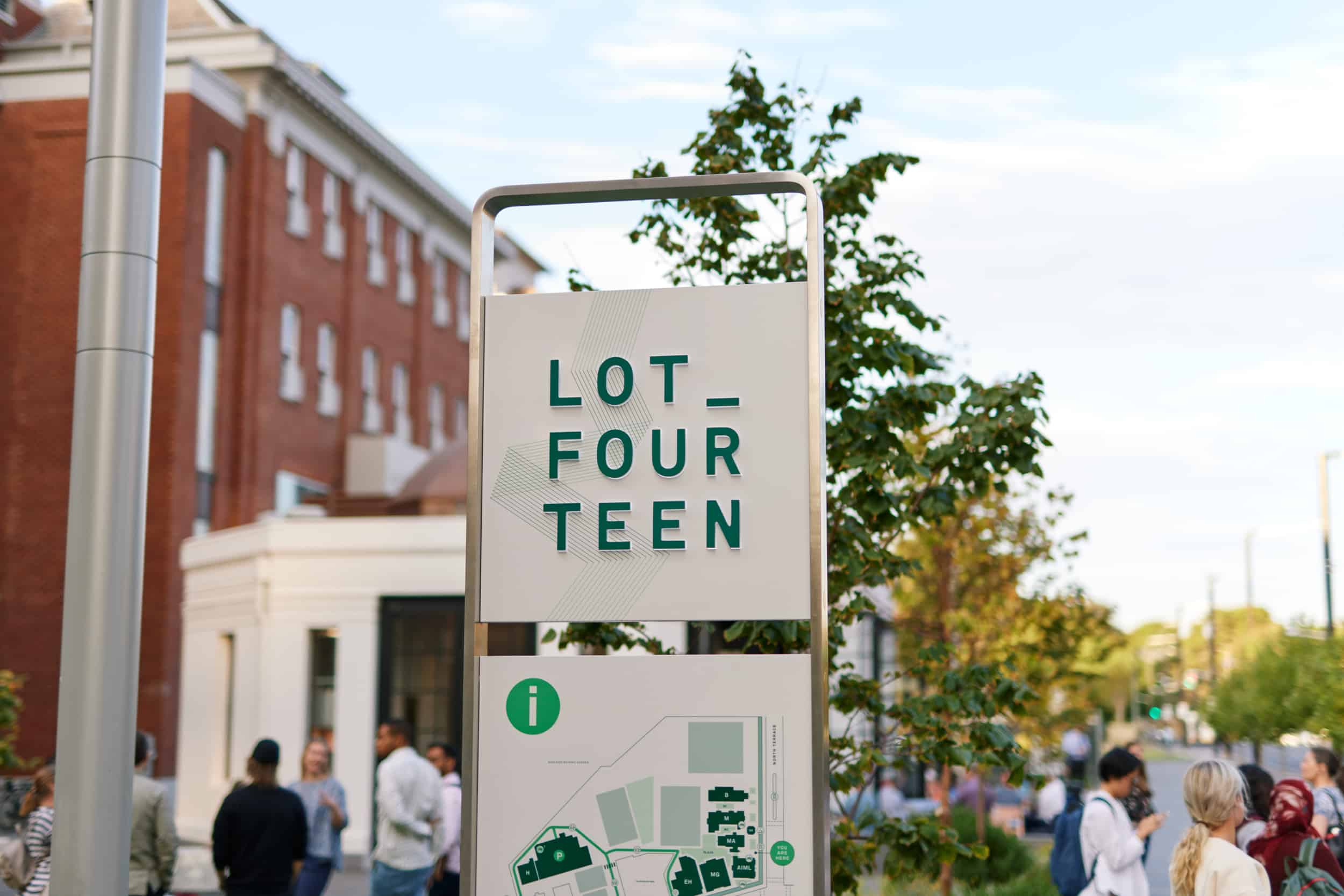 Modern outdoor wayfinding sign reading “LOT FOURTEEN” with an information map below, set in an urban precinct with trees, red brick buildings, and people gathered in the background.