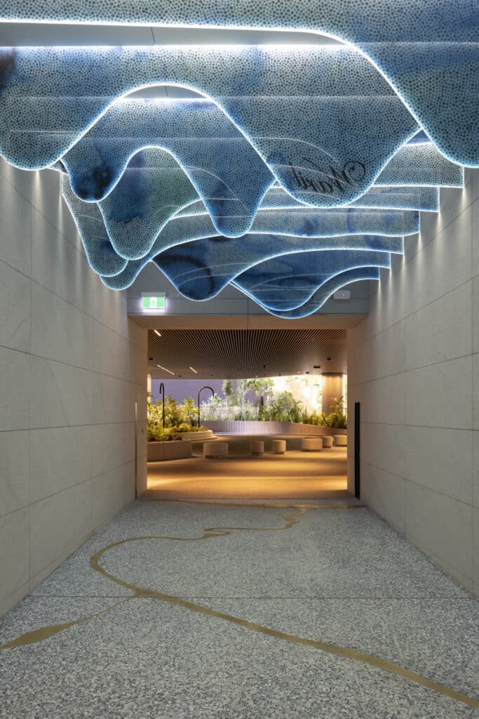 Hallway interior featuring a sculptural blue wave installation on the ceiling, illuminated edges, and a landscaped courtyard with seating beyond.