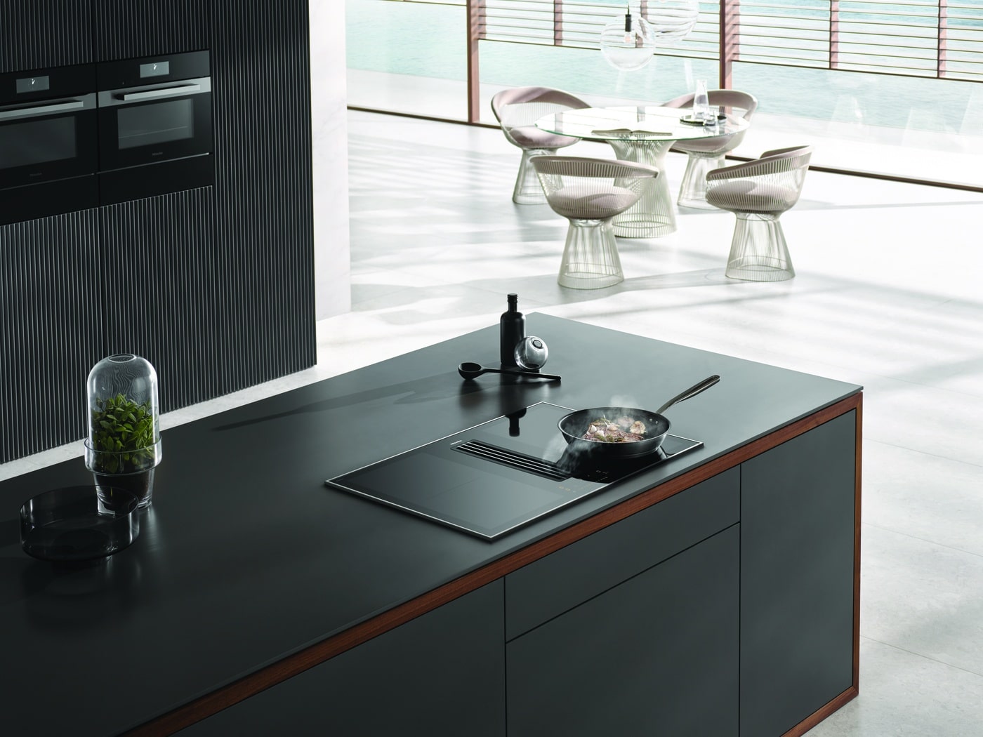 Modern kitchen with a black island bench and integrated cooktop, overlooking a bright dining area with contemporary chairs and large windows.