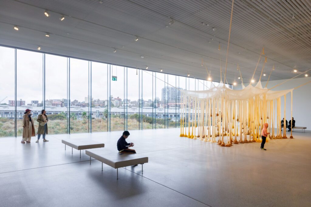 Spacious gallery interior with floor-to-ceiling windows, visitors, and a large hanging textile installation.