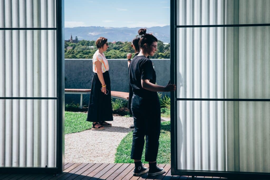 View through open doors to a rooftop garden where three people stand on a path, with greenery and distant hills in the background.