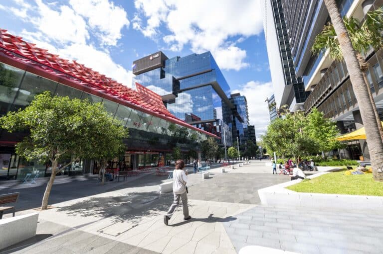 Modern city plaza with glass office towers, a red canopy building, trees, and people walking and relaxing outdoors.