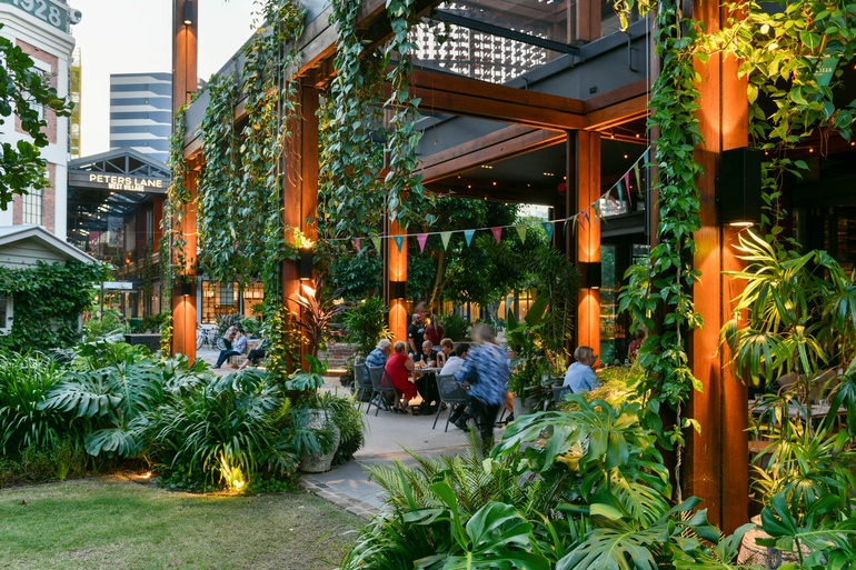 Contemporary outdoor dining courtyard with lush planting, timber structure, and people socialising in urban retail precinct