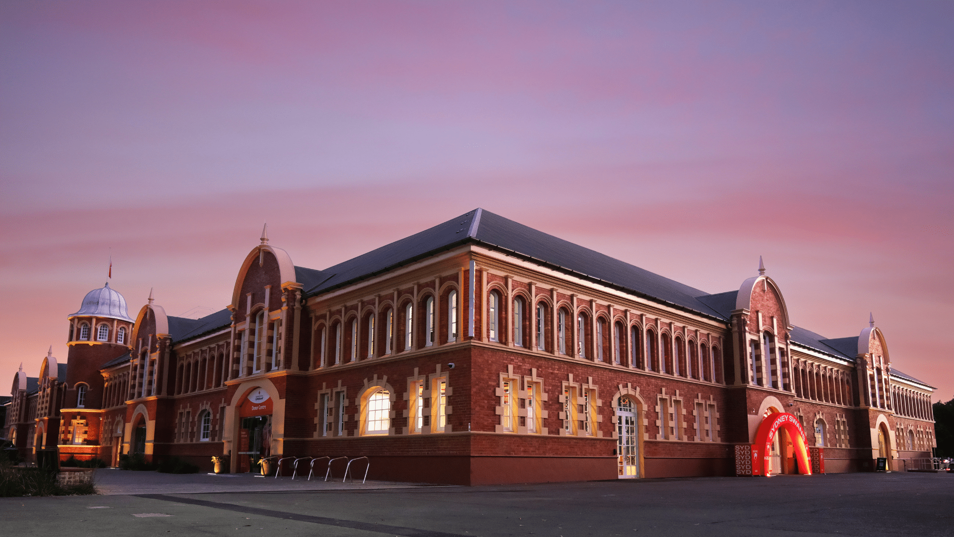 Exterior view of Sydney Swans HQ, a large heritage-style red-brick building with arched windows and decorative detailing, illuminated at dusk under a pink sky, with a glowing red entrance arch and open paved forecourt in the foreground.