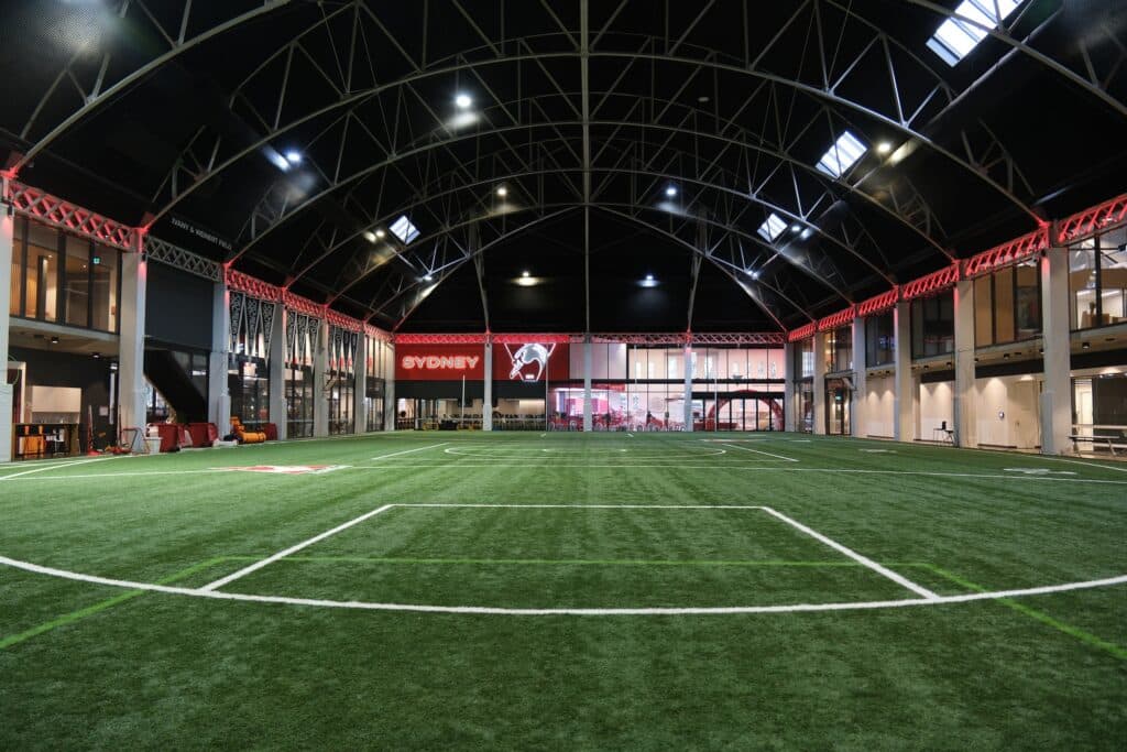 Interior of Sydney Swans HQ featuring a large indoor training field with artificial turf, marked sports lines, and a high arched steel roof, surrounded by glass-walled facilities and illuminated with red accent lighting and “Sydney” team branding.