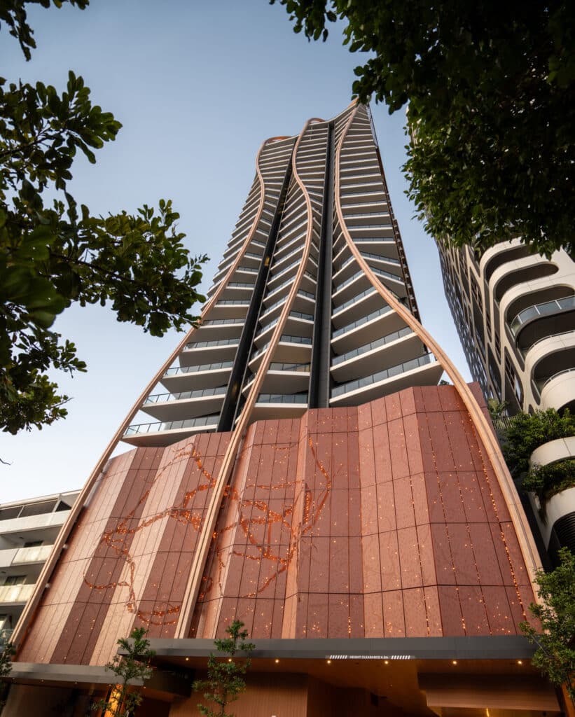 Tall modern residential tower with curved balconies and a copper-toned facade, viewed from below through trees.