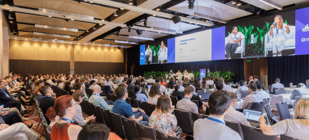 People sitting in the audience at a conference listening to three speakers who are also project on screens