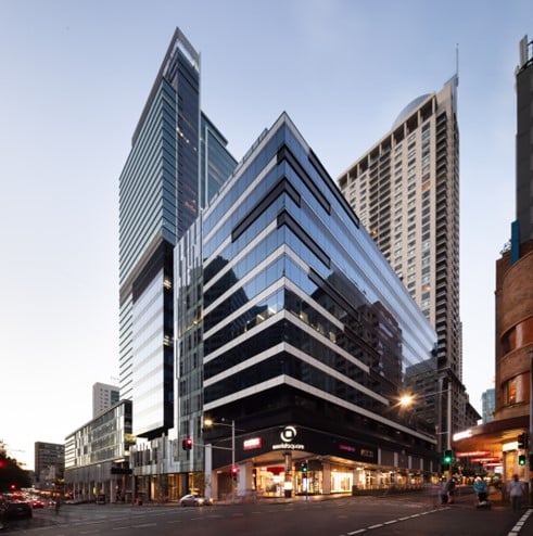 Modern glass office tower at a city intersection, featuring reflective façades, street-level retail, and surrounding high-rise buildings at dusk.