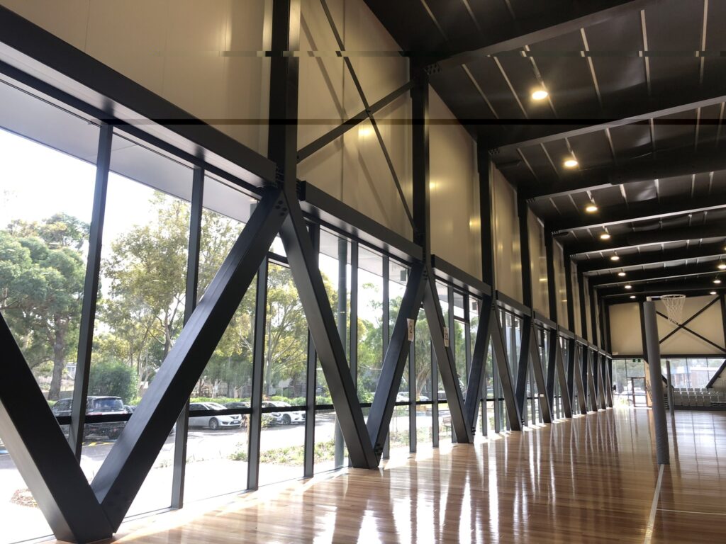 Indoor sports hall with timber floor, tall glass windows, and black steel beams letting in natural light.