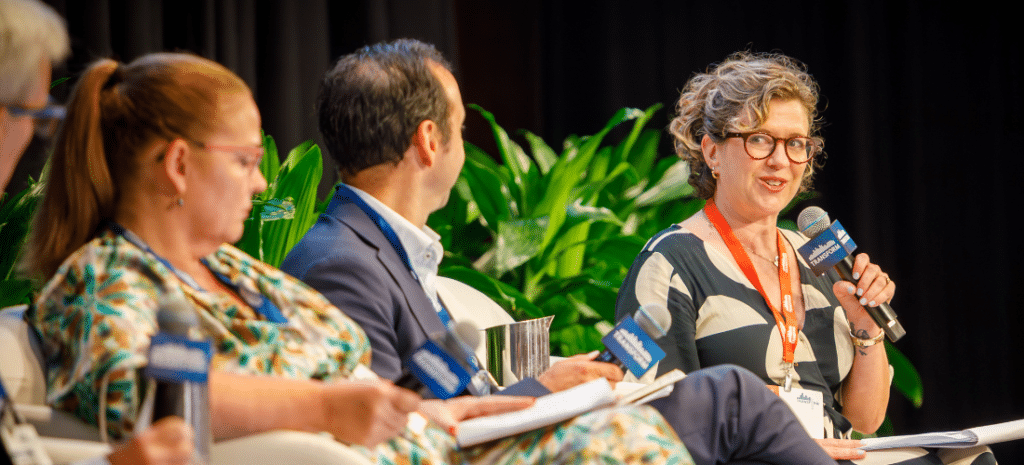 A woman speaking on stage at a conference, looking at the rest of the panel who is seated next to her