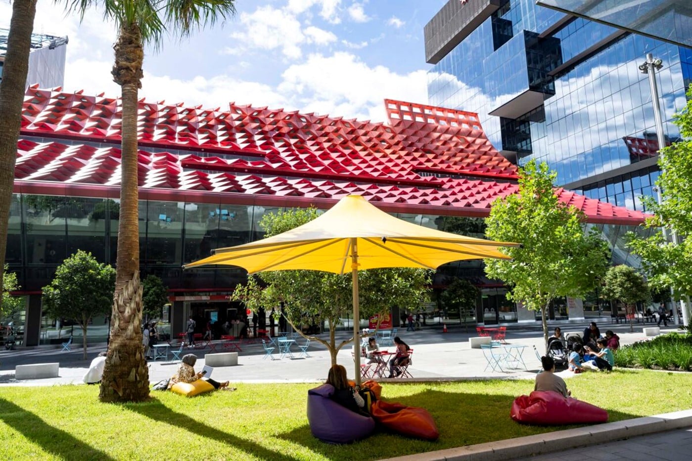 Sunny urban plaza with a red geometric canopy, outdoor seating, and people relaxing on grass under a large umbrella.